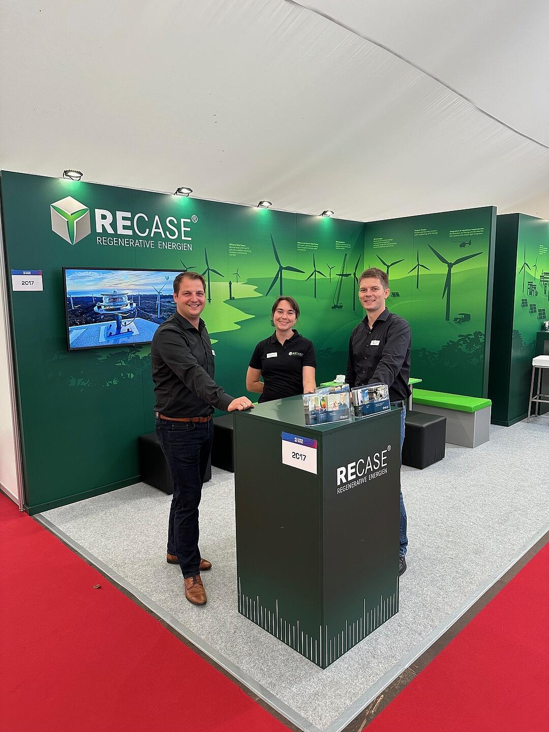 Three RECASE employees stand at the counter of a RECASE trade fair stand.