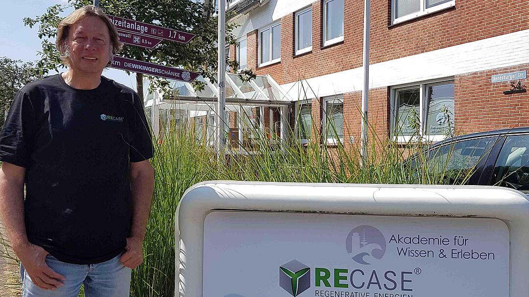 A man in a black T-shirt, jeans and sandals stands next to the Recase company sign.